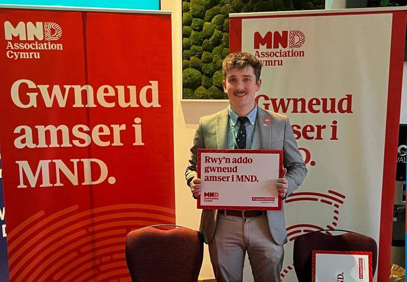 A member of MND Association staff in front of a pull-up banner that says ‘Gwneud amser i MND’.