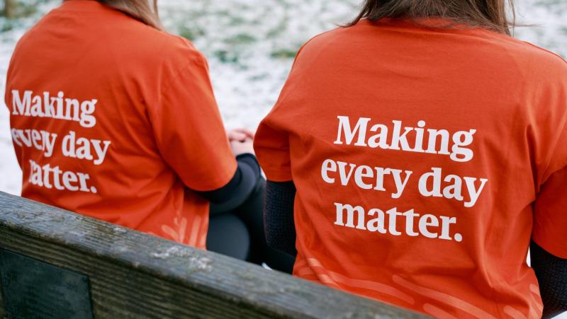 Two people are sitting with their backs to the camera. They are wearing MND Association t-shirts which say 'Making every day matter' on the back.